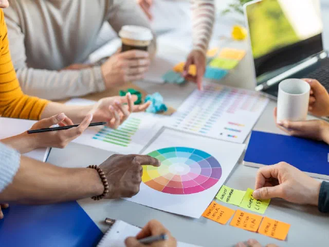 A team around a desk covered in colour wheels, printed colour charts and sticky notes, one hand pointing at a colour wheel