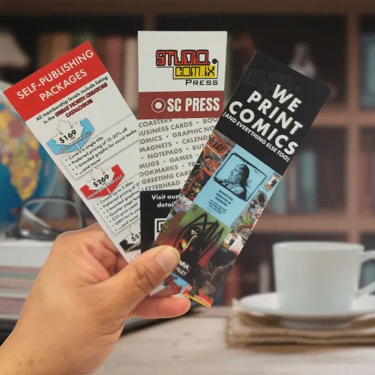 Three SC Press bookmarks fanned in one hand, one reading “We print comics (and everything else too)”