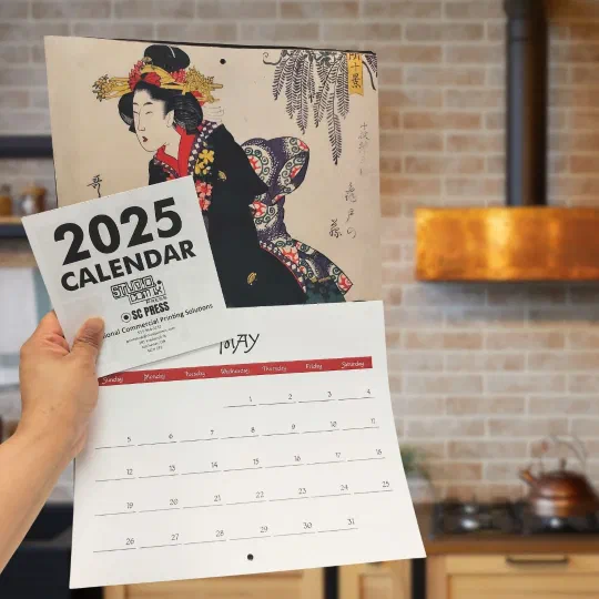 An SC Press 2025 wall calendar held open, an ukiyo-e print above the month grid